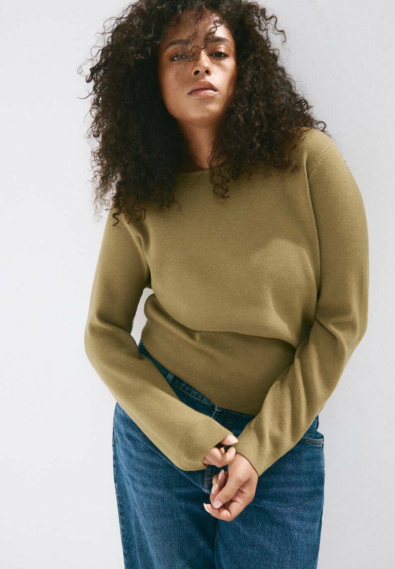 Femme aux cheveux bouclés portant un haut à manches longues vert olive et un jean bleu, debout devant un fond clair uni.