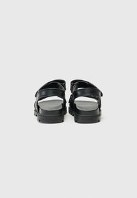 Μαύρα δερμάτινα σανδάλια με ρυθμιζόμενες λουρίδες, επίπεδες σόλες και υφή. Η πίσω όψη δείχνει το ανοικτό σχέδιο στη φτέρνα και στα δάχτυλα.