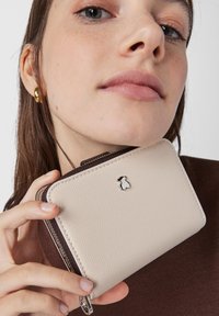 Beige leather wallet with a textured surface and dark brown zipper. Features a silver accent logo on the front. Compact and rectangular shape.