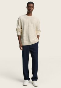 Cremfarbener Strickpullover mit strukturiertem Muster und gerippten Bündchen, kombiniert mit dunkelblauen Hose und weißen Sneakers. Einfaches und modernes Outfit.