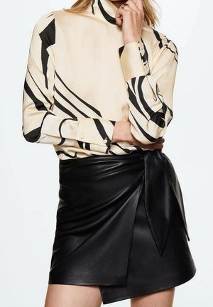 Vrouw draagt een crème blouse met zwarte abstracte strepen en een zwarte asymmetrische leren wikkelrok, poseert met één hand bij haar gezicht.