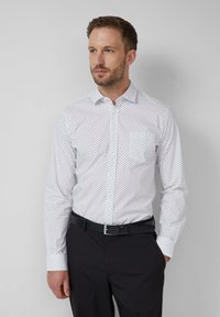 Homme portant une chemise blanche à manches longues avec des pois noirs, une ceinture noire et un pantalon noir, se tenant avec une main dans la poche.