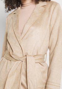 Cappotto avvolgente in suede beige con cintura, tasche laterali e dettagli sul colletto. Presenta una texture liscia e un finish in tessuto morbido.