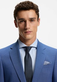 Abito blu con texture, camicia azzurro chiaro, cravatta nera e fazzoletto a quadri nella tasca. L'abito presenta rever a incavo e una vestibilità su misura.