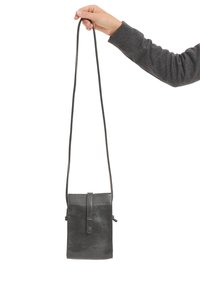 Una mano tiene una borsa a tracolla in pelle nera per il sottile manico. La borsa ha una patta anteriore con chiusura a scatto e un logo embosso discreto.