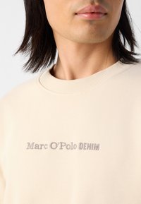 Bežinė sportinė palaidinė su apvaliu kaklu, priešingoje pusėje su dailiu Marc O'Polo DENIM logotipu šviesiai pilka siuvinėjimu. Lygus audinio paviršius.