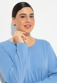 Světle modrá košile s dlouhými rukávy a kulatým výstřihem, jemná textura a volný střih. Modelka má vlasy po ramena a pózuje v uvolněné pozici.