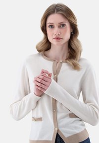 Hellbeige Strickjacke mit beigem Saum, Knopfverschluss vorne und zwei kleinen Taschen. Der Stoff wirkt weich mit einer glatten Textur.