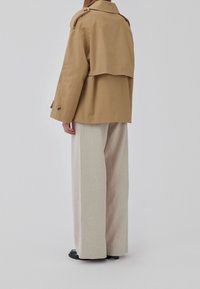 Tan trenchcoat med draperet ryg og to knaplukninger, parret med lysebeige brede bukser og sorte sko.