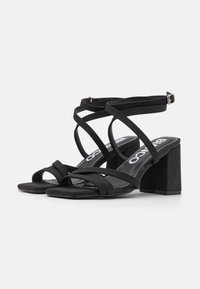 Musta mokkanahkainen paksupohjainen sandaali, jossa on useita ohuita hihnoja ja nilkkahihnan solki, kuvattuna valkoista taustaa vasten.