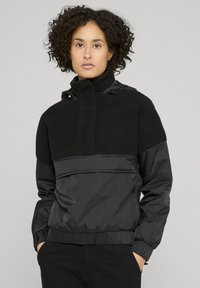 Svart jakke med teksturert fleece øvre del, kontrasterende glatt nedre seksjon, justerbar hette og elastiske mansjetter for en tettsittende passform.