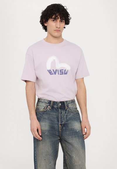 Heled lilla puuvillane T-särk, millel on valgest ja lillast koosnev graafiline "EVISU" logo. Kannan koos kulunud siniste teksapükstega, millel on värviplekid.