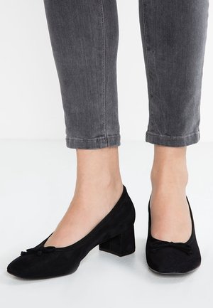 Fødder iført sorte ruskindssko med lav blokhæl og små sløjfer, kombineret med korte mørkegrå jeans.