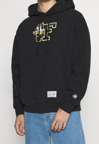 Černá mikina s kapucí s grafikou Felixe Kočky, obsahuje kapuci se stahovací šňůrkou, přední kapsu a logo nášivku na lemu. Volný střih.