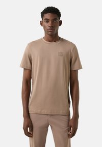 Beige crewneck T-shirt lavet af blød bomuld, med et diskret præget logo på brystet og korte ærmer. Letvægts- og minimalistisk design.