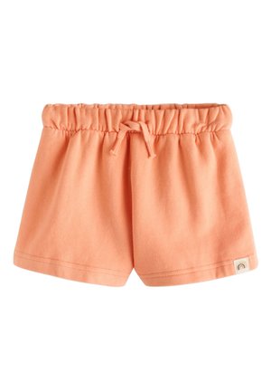 Junges Mädchen mit lockigem Haar, das einen pfirsichfarbenen Hoodie, ein gestreiftes Shirt, passende Shorts, rosa Socken und pastellfarbene Sneakers trägt, steht und lächelt.