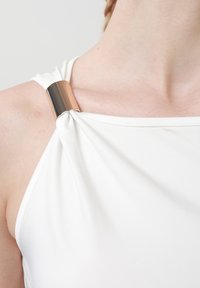 Haut blanc sans manches, froncé et maintenu à l'épaule par une large boucle métallique brillante, laissant apparaître la clavicule et le haut du bras.