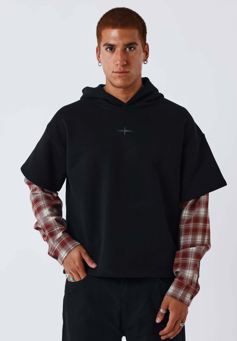 Jeune homme aux cheveux roux courts portant des boucles d'oreilles anneaux, un sweat à capuche noir à manches courtes par-dessus un t-shirt à manches longues à carreaux rouges, et un pantalon noir, debout devant un fond blanc.