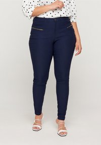Navyblauwe skinny jeans met twee met rits afgesloten voorzakken, gemaakt van stretchdenim met een soepele textuur, gecombineerd met een witte top met stippen.