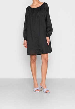 Femme portant une mini robe noire à manches longues ample, associée à des sandales à talons hauts tressées gris clair, debout sur un sol uni.