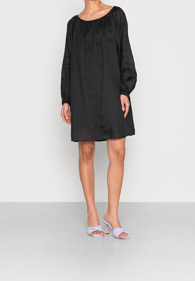 Femme portant une mini robe noire à manches longues ample, associée à des sandales à talons hauts tressées gris clair, debout sur un sol uni.