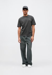 Hombre con gorra gris oscuro, camiseta gris oscuro, pantalones vaqueros holgados oscuros y zapatillas blancas, de pie contra un fondo claro liso.