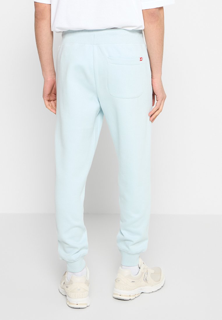 Un pantalon de survêtement bleu clair avec des poignets côtelés, une taille élastique et une poche arrière unique. Présente un petit logo rouge au-dessus de la poche.