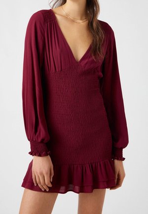 Robe mini bordeaux avec de longues manches bouffantes, un décolleté en V profond et un corsage smocké. Présente un ourlet à volants et un tissu doux et texturé.