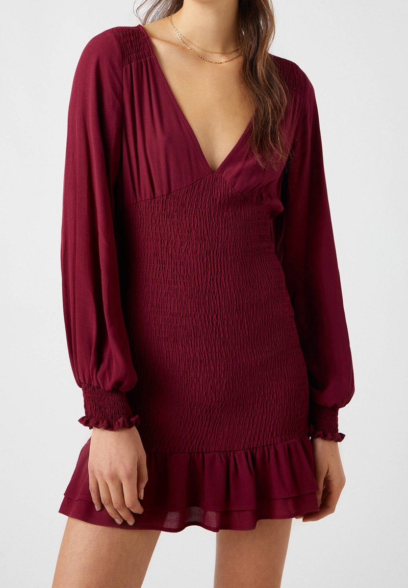 Vestido mini burdeos con mangas largas y abullonadas, escote en V profundo y cuerpo fruncido. Presenta un dobladillo con volantes y una tela suave y texturizada.