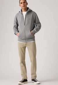 Sudadera gris con cremallera de algodón, que cuenta con un bolsillo tipo canguro y capucha con cordón, combinada con pantalones beige claros y zapatillas bajas negras.