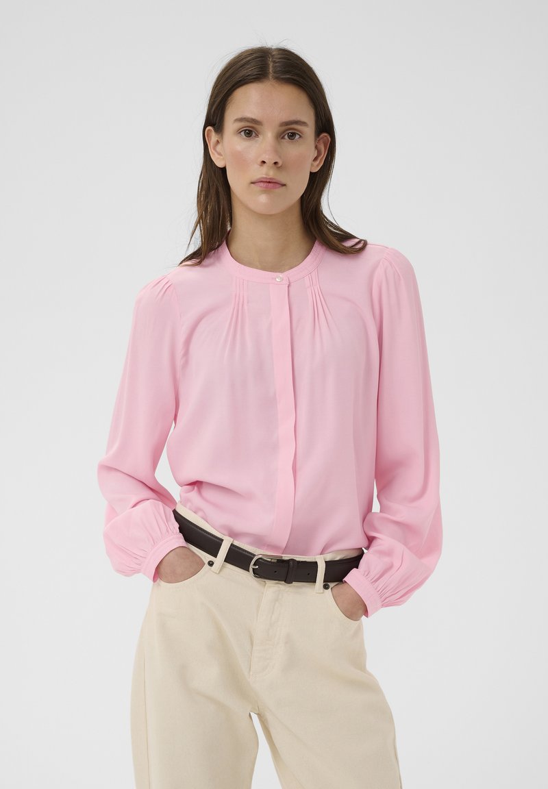 Femme portant un chemisier rose à manches longues rentré dans un pantalon beige avec une ceinture noire, debout les mains dans les poches devant un fond uni.