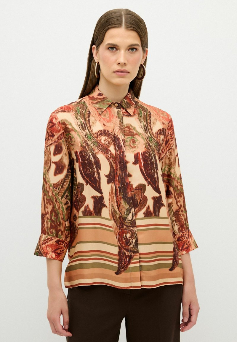 Blouse en satin avec des motifs paisley et rayés dans des tons chauds d'orange, de brun et de vert. Elle possède un col et des manches trois-quarts.