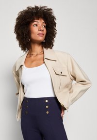 Beige cropped jasje met een puntige kraag, voorzak en gouden rits. Draagt over een wit geribbeld tanktop en marineblauwe broek met gouden knopen.
