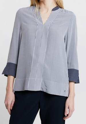 Femme portant un blouse grise avec de fines rayures verticales, col en V et poignets rayés navy, accompagnée de pantalons noirs.