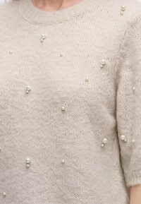 Gros plan d'un pull en tricot couleur crème avec de petits groupes décoratifs de perles sur le devant et les manches.