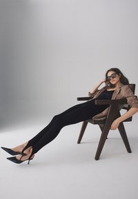 Mustast keha kujundav kombinesoon, kontsakingad lõigetega detailidega, helepruun suur jakk, puidust tool, neutraalne taust.