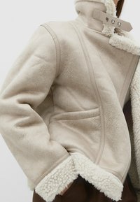 Béžová bunda z beránkoviny s vysokým límcem, kontrastním podšívkou a zapínáním na zip. Má prošívané detaily a měkkou strukturu na manžetách a lemu.