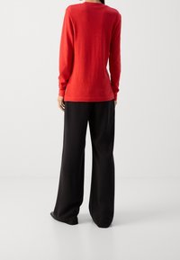 Top rosso a maniche lunghe con scollatura a girocollo abbinato a pantaloni neri a gamba larga; il tessuto appare liscio e leggero.