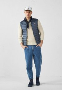 Gilet matelassé bleu marine sur un pull crème, jeans bleu clair et chaussures foncées. Casquette de baseball blanche avec logo. Écharpe bleu foncé autour du cou.