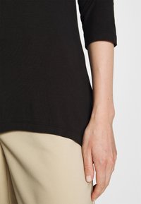 Svart, mjukt tyg med trekvartslånga ärmar, med en rundad nederkant; kombinerat med beige, texturerade byxor. Synlig hand på tyget.