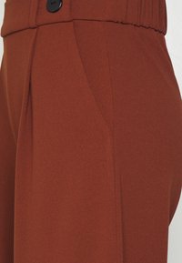 Pantalons couleur rouille en tissu lisse, dotés d'un seul bouton noir et de poches latérales avec un design ajusté.