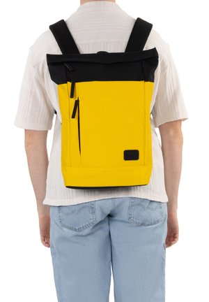 Personne portant un sac à dos jaune et noir avec deux fermetures éclair verticales à l'arrière, habillée d'une chemise blanche à manches courtes et d'un jean bleu clair.
