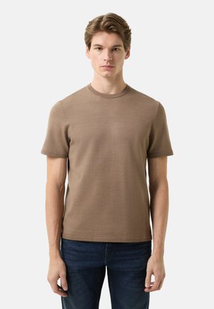 Junger Mann, der ein eng anliegendes, kurzärmeliges braunes Strickhemd und dunkelblaue Jeans trägt, vor einem einfarbigen hellen Hintergrund steht.