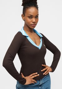 Νεαρή γυναίκα με τα χέρια της δεμένα στη μέση, φοράει μακρυμάνικο καφέ μπλουζάκι με γιακά με μπλε ρίγες και μπλε τζιν, πάνω σε ένα απλό φωτεινό φόντο.