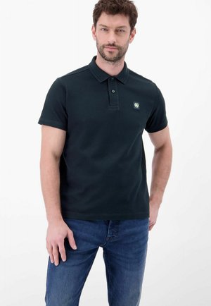Mann trägt ein schwarzes kurzärmeliges Polohemd mit kleinem Brustlogo und blaue Jeans, steht vor weißem Hintergrund und blickt nach vorne.