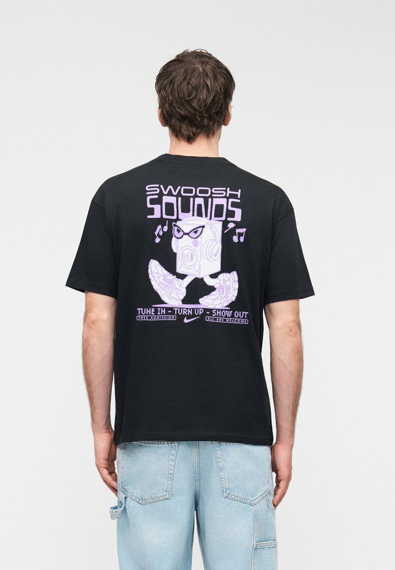 Homme portant un t-shirt noir avec un graphisme violet représentant un haut-parleur ambulant portant des lunettes et des baskets, avec le texte "Swoosh Sounds" et "Accordez-vous - Montez le son - Exprimez-vous."