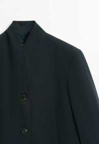 Svart blazer med minimalistisk krage och två synliga mörka knappar framtill mot en vit bakgrund.