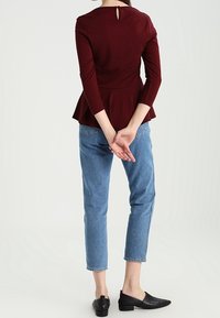 Bordeauxfarbener Peplum-Top mit einem Schlüsselloch auf der Rückseite, kombiniert mit hellblauen, umgeschlagenen Jeans und schwarzen Slippern. Glatter Stoff, figurschmeichelndes Design.
