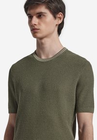 Kortärmad, olivgrön stickad skjorta med ett strukturerat mönster och kontrasterande krage. Har en rundad halsringning och åtsittande ärmar.