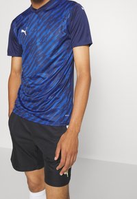 Marinblå träningströja med vinklade blå mönster, korta ärmar och v-ringning. Tillsammans med svarta shorts och vita strumpor.